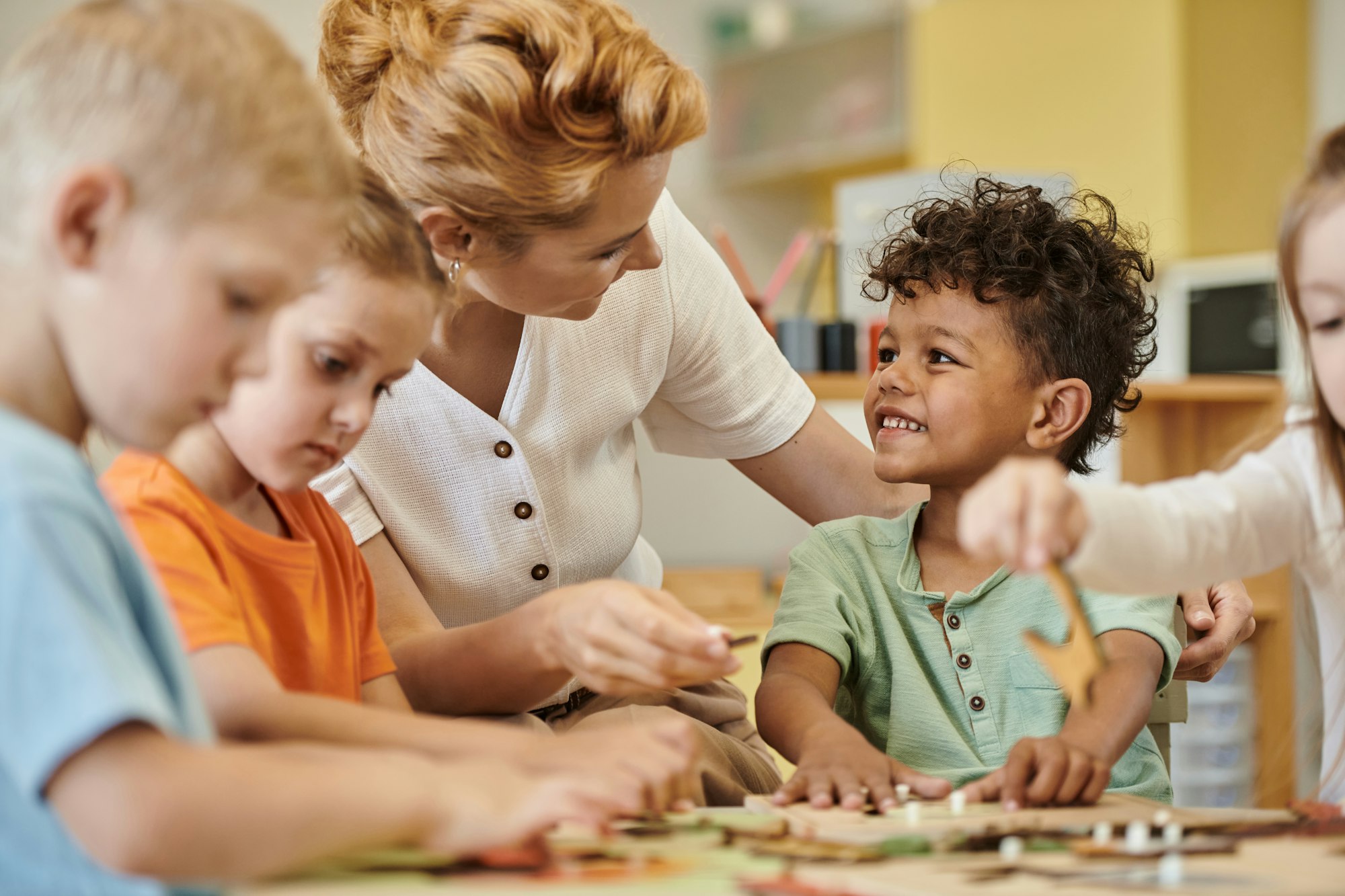 teacher talking to smiling african american boy while kids playing in montessori school